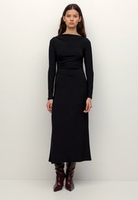 Fiatal nő áll hosszú ujjú, fekete midi ruhában, amely szűkített derékrésszel rendelkezik, és bordó magassarkú csizmát visel, egyszerű világos háttér előtt.