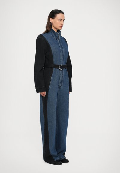 Salopette in denim con un design bicolore composto da sezioni blu e nere, colletto alto, zip frontale e una cintura aderente in vita.