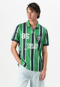 Young man wearing green, black, and white vertical striped polo shirt with "85" and "tommy jeans" text, paired with light blue jeans, standing.