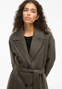 Oliivivihreä villasekoitekappale, jossa on tikkupielet, yksirintainen napitus ja yhteensopiva vyö sidottuna vyötärölle.