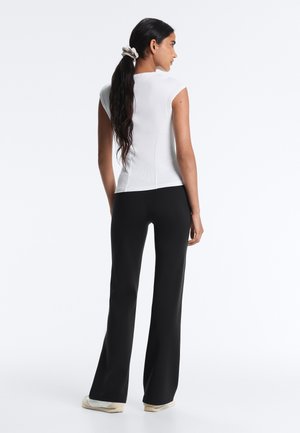 Donna con lunghi capelli scuri legati con uno scrunchie, che indossa un top bianco senza maniche, pantaloni neri svasati e sneakers bianche, in piedi di spalle.