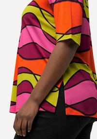 Bunt bedrucktes Kurzarmoberteil mit kräftigen, wellenförmigen Mustern in Orange, Pink, Grün und Schwarz. Hergestellt aus einem glatten Stoff mit seitlichen Schlitzen.