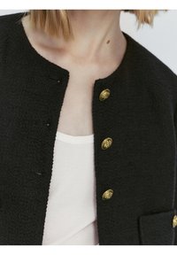 Svart texturerad jacka med rund halsringning, tre gyllene dekorativa knappar och en framficka. Buren över en ljusfärgad topp.