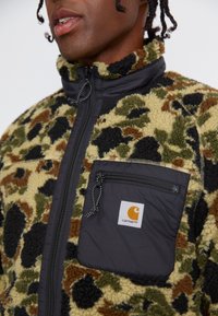 Veste en polaire avec une poche avant noire, ornée d'un motif camouflage dans des tons de vert olive, marron et beige. Fermeture éclair et col montant.