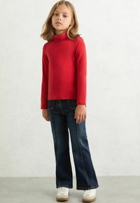 Maglione rosso a maniche lunghe con collo alto abbinato a jeans in denim scuro a gamba svasata e scarpe da ginnastica bianche, caratterizzato da una texture liscia e un design semplice.