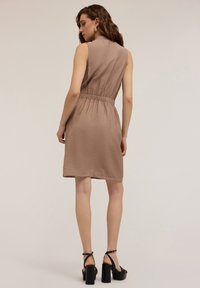 Rochie scurtă, fără mâneci, de culoare marron, cu un material texturat, talie strânsă și tiv la genunchi. Asortată cu sandale negre cu toc gros.