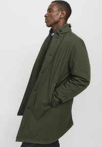 Cappotto verde oliva di media lunghezza con design strutturato, caratterizzato da un alto colletto, chiusura frontale a bottoni nascosta e tasche laterali. Texture liscia.