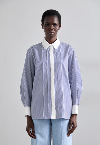 Chemise à rayures bleues et blanches avec un col double blanc, à manches longues et coupe droite. Portée avec un pantalon en denim bleu clair.