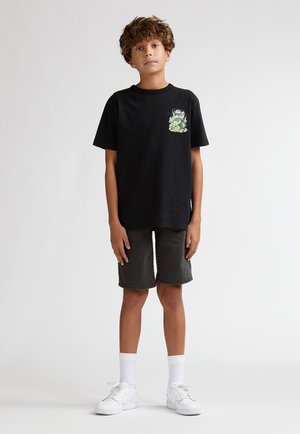 Tricou negru din bumbac cu un imprimeu frontal, asortat cu pantaloni scurți din denim gri închis. Modelul poartă adidași albi și șosete albe până la glezne.