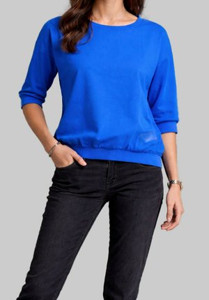 Femme portant un haut bleu vif à manches longues avec manches retroussées et un jean noir, debout avec une main dans la poche sur un fond gris.