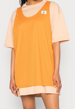 Kvinde iført oversized beige t-shirt under løs orange mesh ærmeløs sportsjersey med lille logo-patch nær skulderen.