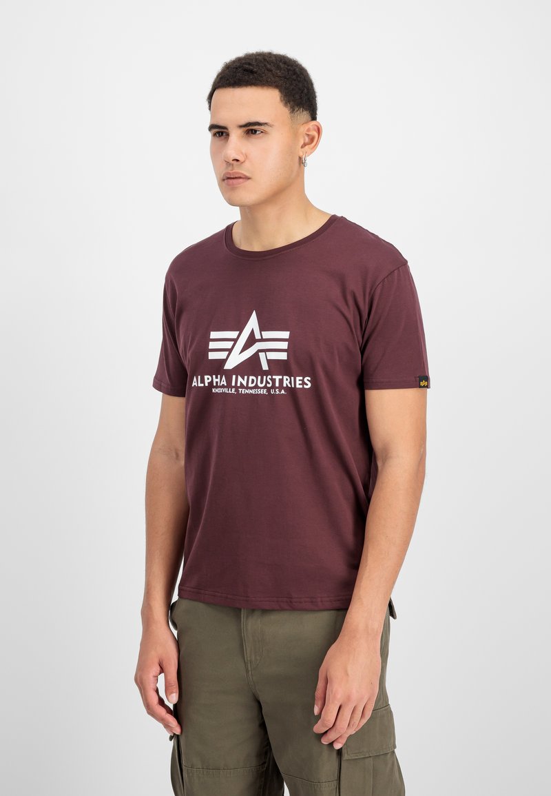 Burgunderfarbenes Baumwoll-T-Shirt mit einem weißen Logo und Design sowie drei horizontalen Streifen, kombiniert mit olivgrünen Cargo-Hosen.