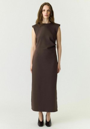 Femme aux longs cheveux bruns, vêtue d'une robe midi sans manches de couleur marron foncé et de chaussures noires à bouts pointus, debout devant un fond clair uni.