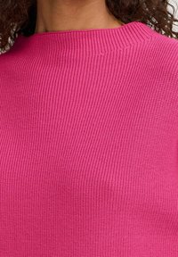 Pull en maille rose ajusté avec une texture côtelée et un col montant, présentant un léger éclat. Vue en gros plan centrée sur la zone de la poitrine.