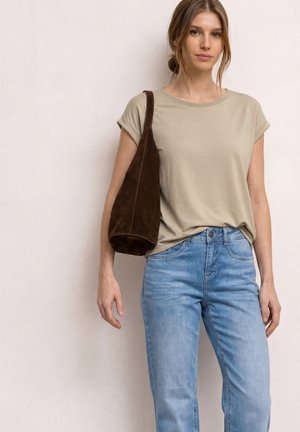 Młoda kobieta ubrana w jasnobeżowy top z krótkim rękawem, jasnoniebieskie jeansy, niosąca ciemnobrązową torebkę na ramię, stoi na tle jednolitej ściany.