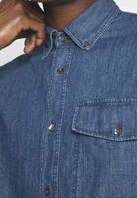 Camicia di denim blu scuro con colletto button-down, tasca frontale e bottoni argentati. Il tessuto ha un aspetto leggermente texturizzato e intrecciato.