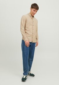 Beige Hemd mit Knöpfen und Tasche, kombiniert mit lockeren blauen Jeans und grünen Sneakers mit weißen Akzenten. Lässige Passform mit strukturierter Oberfläche.