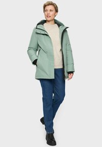 Vrouw in een lichtgroene gewatteerde winterjack, beige trui, blauwe spijkerbroek en zwarte laarzen staat met haar handen in de zakken.
