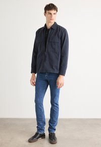 Chaqueta navy con cremallera y dos bolsillos en el pecho, llevada sobre una camiseta polo negra, combinada con jeans azules y zapatillas negras en un fondo neutro.