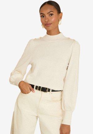 Femme portant un pull crème avec des boutons dorés sur les épaules et un pantalon taille haute assorti, accessoirisée d'une ceinture noire et de boucles d'oreilles dorées.