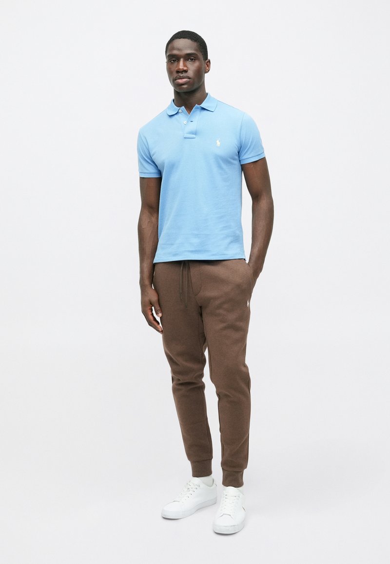 Hellblaues Poloshirt mit kurzen Ärmeln, kombiniert mit braunen Jogginghosen. Das Modell trägt weiße Sneaker. Einfaches Design mit minimaler Markenkennzeichnung.