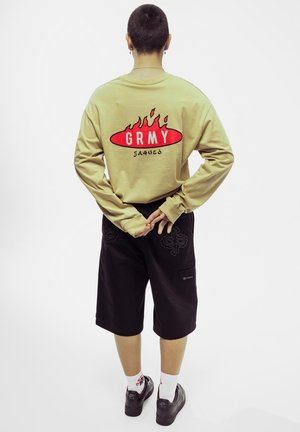 Camiseta de manga larga color caqui con un gráfico de llamas rojas y el texto "GRMY" en la parte posterior, combinada con shorts negros que presentan un bordado intrincado.
