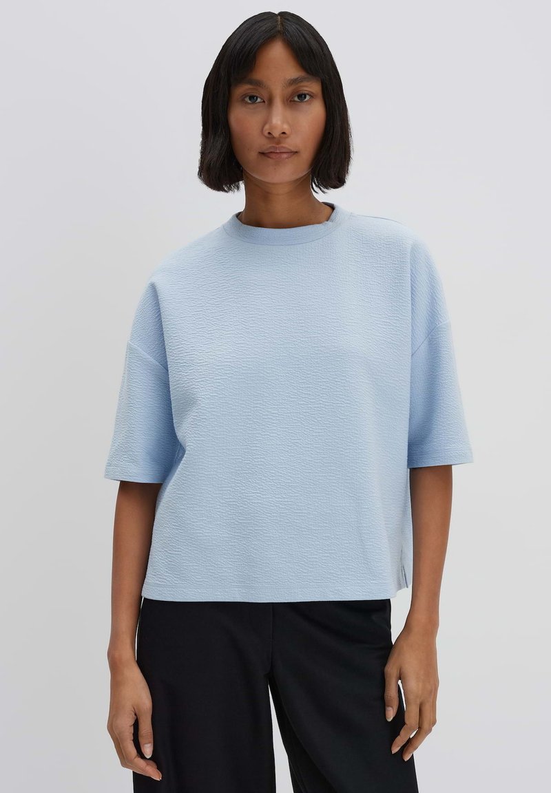 Lichtblauw oversized shirt met korte mouwen van een gestructureerde stof, ronde halslijn en zijsplitten, gecombineerd met zwarte broek. Eenvoudig ontwerp.