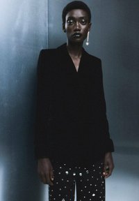 Blazer de terciopelo negro con solapas de chal, combinado con pantalones negros con tachuelas plateadas. Fondo en tonos gris suaves.