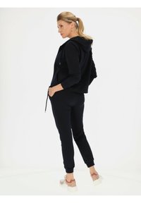 Czarna bluza z kapturem na zamek i sznurkiem, zestawiona z dopasowanymi dresami. Gładki materiał, ściągacze oraz wyraźne sportowe sneakersy.