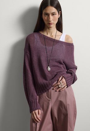 Donna che indossa un maglione lavorato a maglia violaceo largo sopra una maglietta bianca, pantaloni malva, una lunga collana e anelli moderni in argento, in piedi contro una parete grigia chiara.