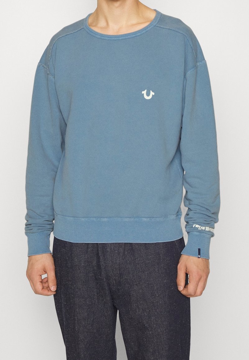 Homme portant un sweat-shirt bleu délavé à manches longues avec un petit logo blanc sur la poitrine et un pantalon sombre, sur un fond uni.