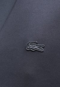Tissu gris foncé avec un petit logo de crocodile brodé en fil gris foncé assorti, centré sur la surface.