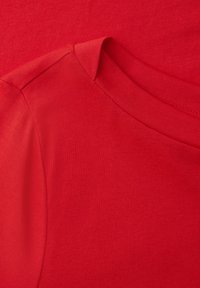Gros plan d'un t-shirt rouge à col rond montrant l'encolure et la couture de l'épaule avec une texture de tissu lisse et finement tricotée.