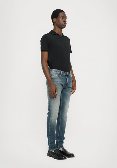 Črna polo majica, svetlo do temno modri kavbojki z rahlim beljenjem, črne čevlje. Model stoji pokončno ob beli steni.