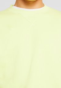 Sudadera amarilla claro con cuello redondo, que presenta una textura suave y un detalle de costura mínimo. La tela parece suave y casual.