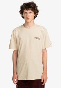 Beige T-shirt met korte mouwen, gemaakt van katoen, met een klein bruin logo op de borst en aanvullende logo-tekst op de mouw.