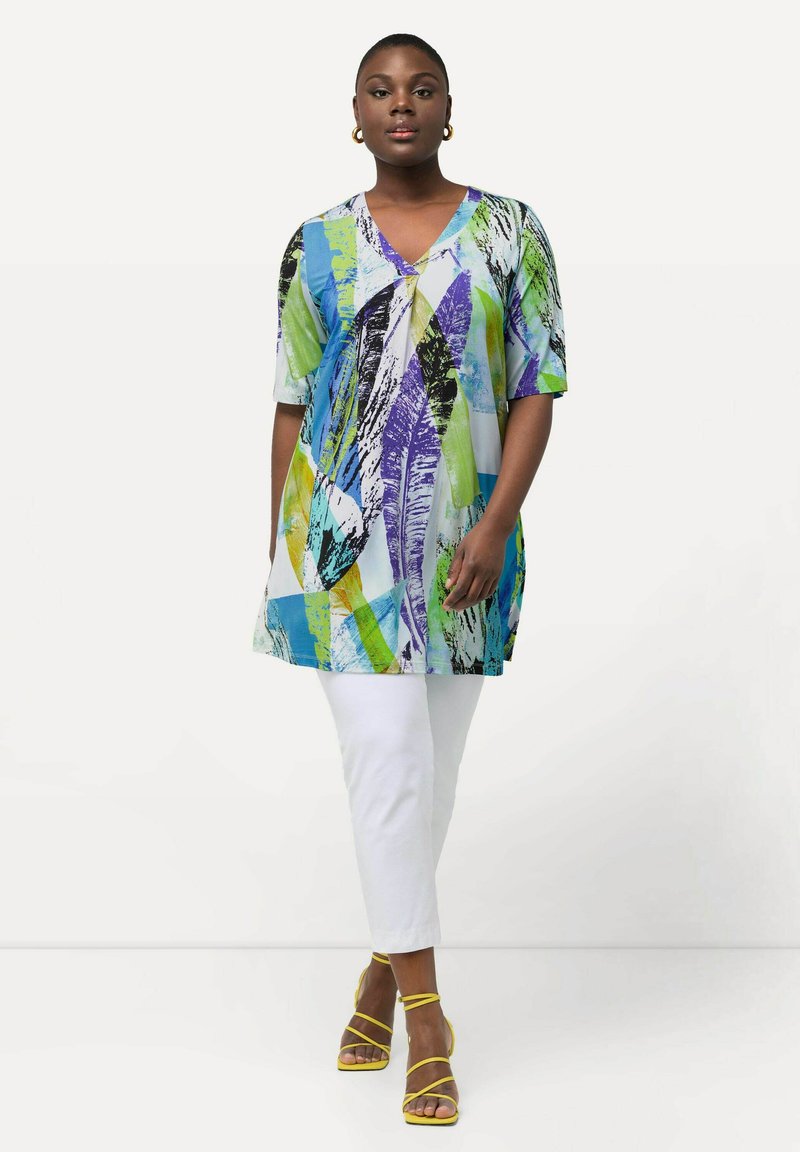 Túnica multicolor con escote en V, que presenta vibrantes patrones abstractos en azul, verde y púrpura; combinada con pantalones blancos y sandalias amarillas de tiras.