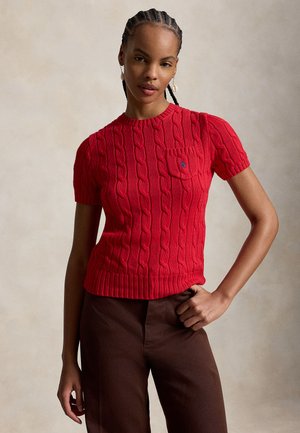 Γυναίκα που φοράει κόκκινο πουλόβερ με κοντά μανίκια και πλέξη κοτσίδα, με τσέπη στο στήθος και καφέ παντελόνι, στέκεται με το ένα χέρι στη μέση μπροστά σε ουδέτερο φόντο.