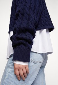 Main portant une manche de pull en maille torsadée bleu marine sur une chemise blanche, reposant près de la poche d’un jean en denim bleu clair.