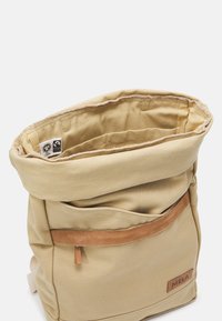 Tan roll-top rugzak gemaakt van canvas, met een leren accent op het voorvak en een logolabel. Het interieur heeft een gevoerd textuur.