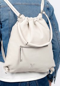 Sac à dos à cordon de serrage perforé blanc avec une texture lisse, poche zippée argentée et détails de bandoulière réglables. Branding discret sur le côté.