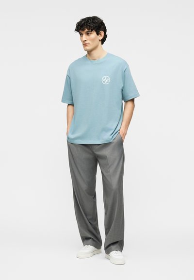 Νέος άνδρας με σγουρά μαλλιά που φοράει ανοιχτό μπλε φαρδιά μπλούζα, γκρι παντελόνια με φαρδιά μπατζάκια και λευκά αθλητικά παπούτσια, στέκεται με τα χέρια στις τσέπες.