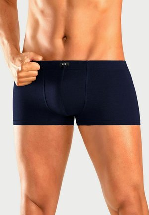 Mann trägt marineblaue Boxershorts, hält den Bund mit einer Hand, zeigt definierten Bauch und obere Oberschenkel vor einfachem Hintergrund.