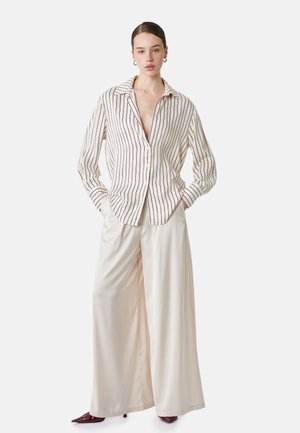 Kvinde iført en beige og brun vertikalt striped knap-skjorte med vide cremefarvede bukser og bourgogne spidse sko, hænderne i lommerne.