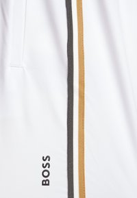 Shorts blancs ornés d'un logo noir et de deux bandes verticales sur les côtés en gris et or. Tissu lisse avec un design épuré.