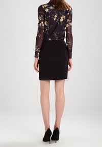 Jupe crayon noire associée à un haut transparent à manches longues avec un motif floral, présentant des fleurs jaunes et blanches sur un fond sombre.