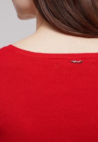 Maglione a costine rosso con scollo rotondo, caratterizzato da un piccolo dettaglio con logo nero nella parte posteriore dello scollo. Texture liscia.