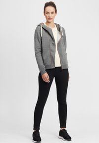 Siva zip-up dukserica s vezicama, uparena s svijetlom majicom i crnim uskim trapericama. Crne sportske tenisice upotpunjuju outfit.
