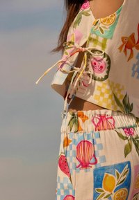 Twee-delig outfit bestaande uit een cropped top en broek met elastische taille. Kleurrijke patronen met fruit, bloemen en lantaarns op een crèmekleurige achtergrond.