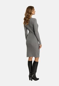 Graues geripptes Strickkleid mit langen Ärmeln, figurbetonter Silhouette, knielang, mit seitlichen Knopfakzenten. Kombiniert mit knielangen schwarzen Stiefeln mit Absatz.